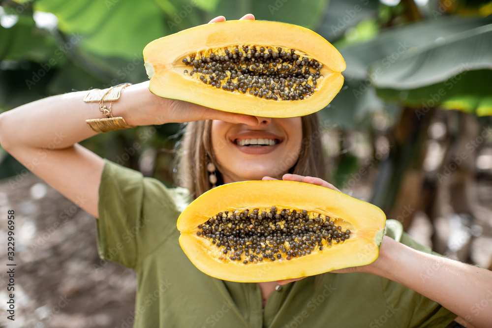 Closeup portrait of a young woman hiding her face behind sliced papaya fruit outdoors on the green b