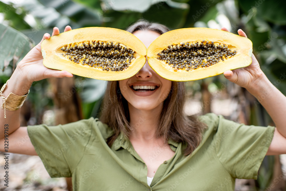 Closeup portrait of a young smiling woman hiding her eyes behind sliced papaya fruit on the green ba