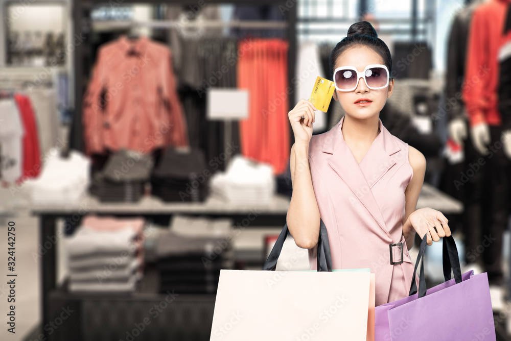 Young pretty cute Asian woman holding credit card and shopping bags with smiley face standing in clo