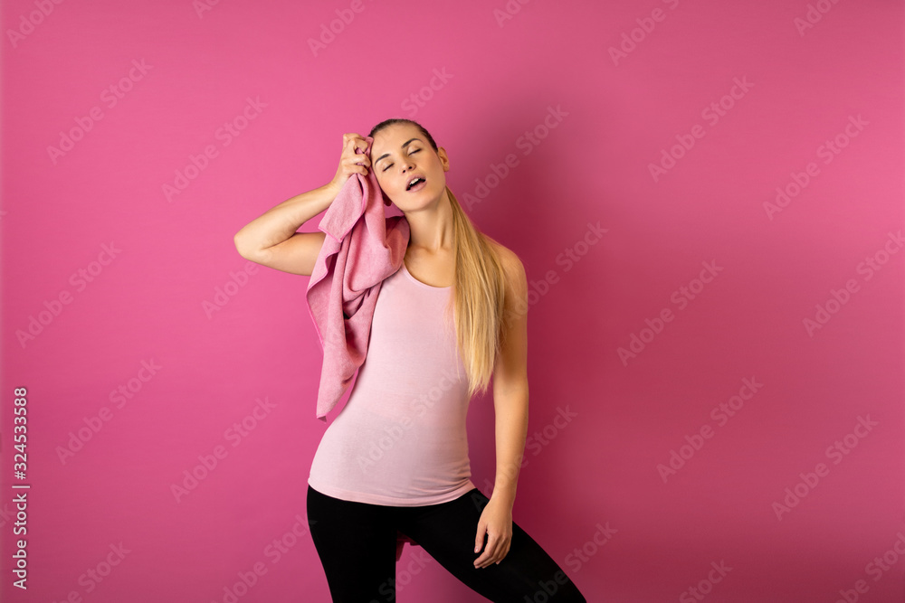 Young fit woman tired after workout with towel on her shoulders on pink background