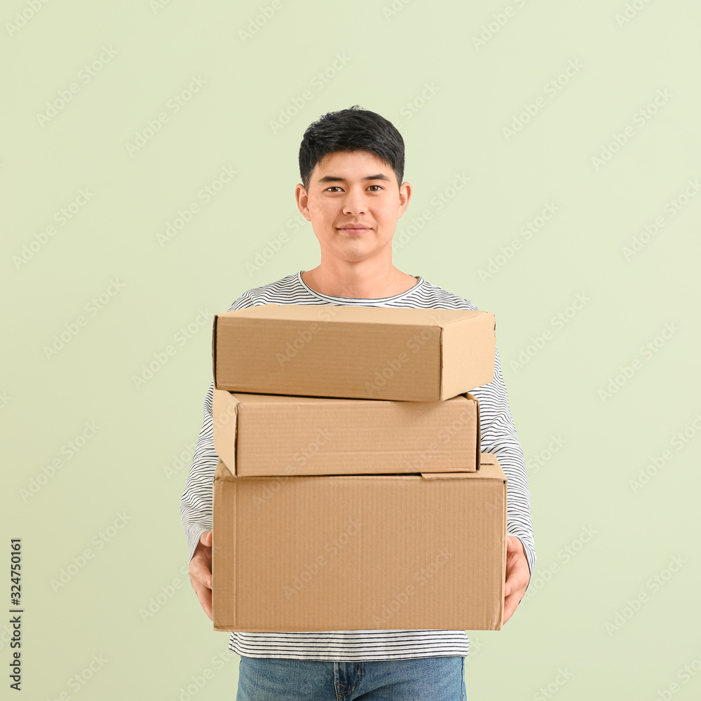 Asian man with moving boxes on color background