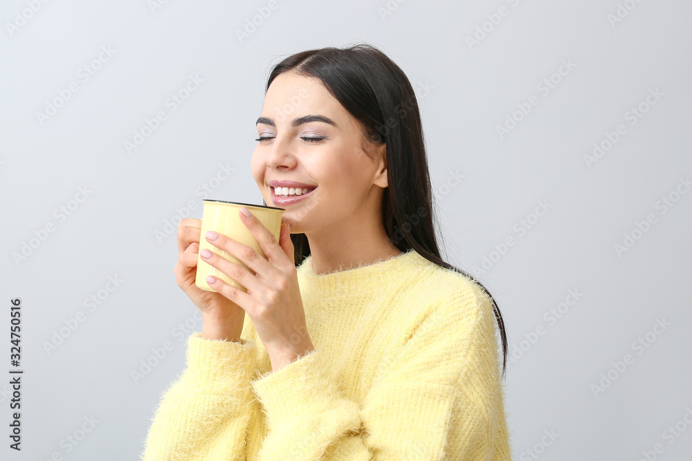 Beautiful young woman in warm sweater drinking tea on light background