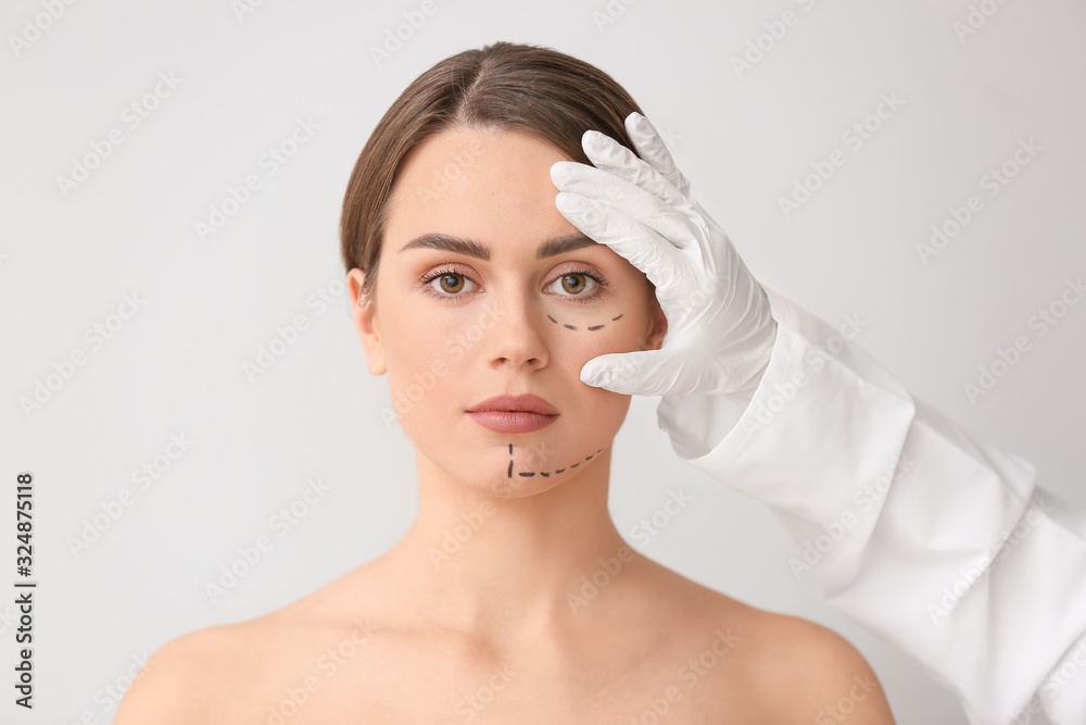 Plastic surgeon touching face of young woman on light background