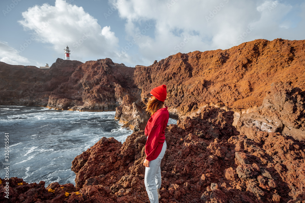 Woman enjoying great views on the rocky ocean coast and a lighthouse on the background, traveling on