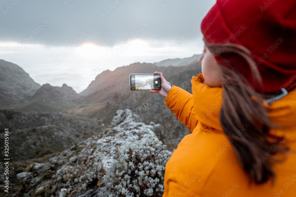 Young woman dressed in bright jacket and hat photographing on phone breathtaking views on the mounta