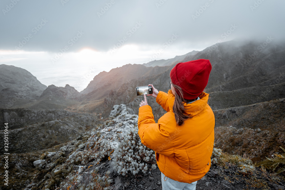 Young woman dressed in bright jacket and hat photographing on phone breathtaking views on the mounta