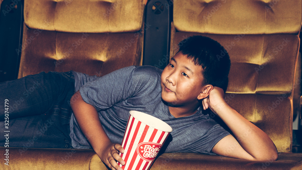 The boy is laying down watching movies and eating popcorn happily and enjoying in the cinema.