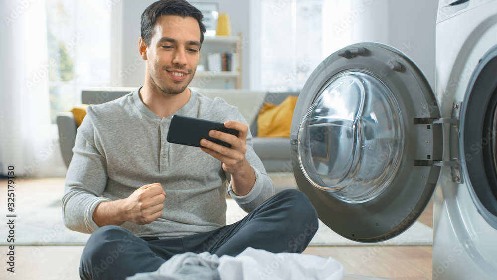 Handsome Smiling Young Man in Grey Jeans Sits in Front of a Washing Machine, Plays Games on His Smar