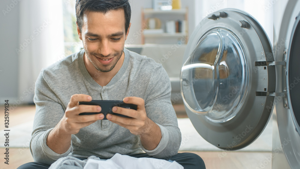 Handsome Smiling Young Man in Grey Jeans and Coat Sits in Front of Washing Machine and Plays Games o