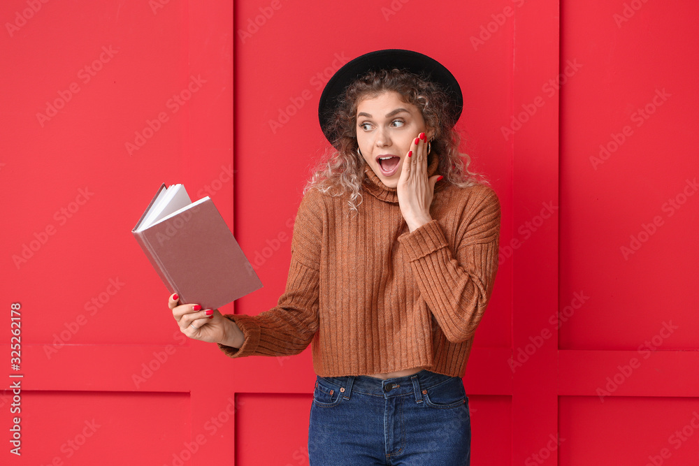 Surprised young woman in warm sweater and with book on color background