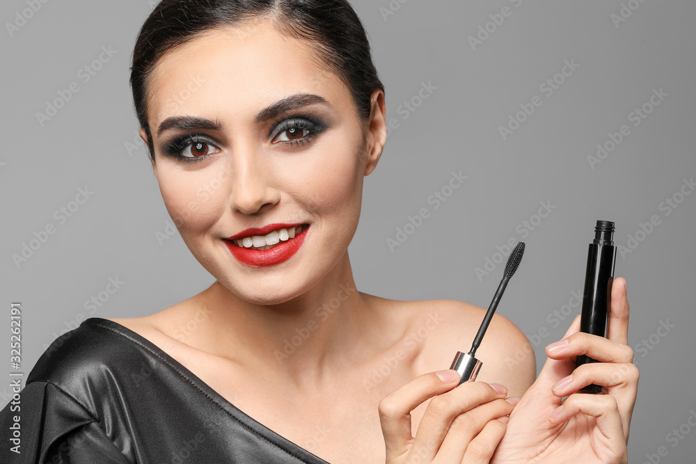 Beautiful young woman applying makeup against grey background