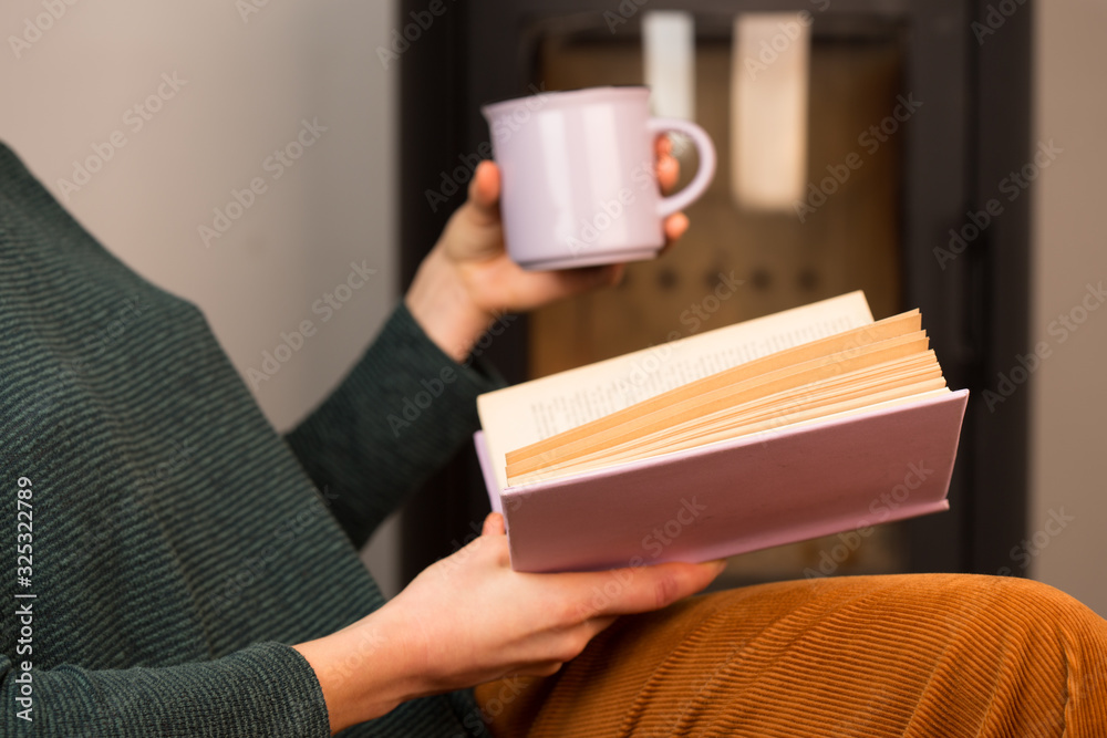 年轻女子坐在壁炉旁的椅子上看书，手里拿着一杯茶