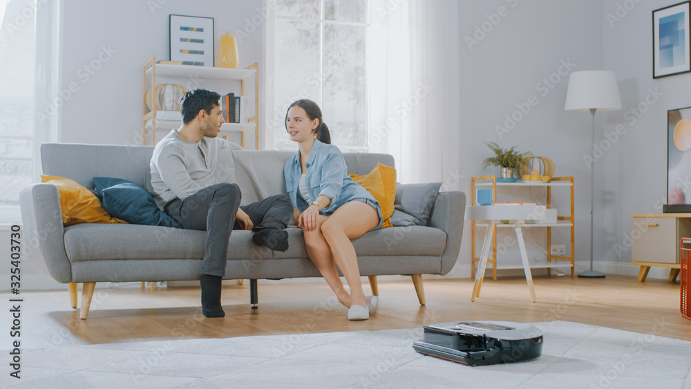 Smart Robot Vacuum Cleaner Sucking Up Dust from a Carpet. Beautiful Couple is Sitting on a Sofa and 