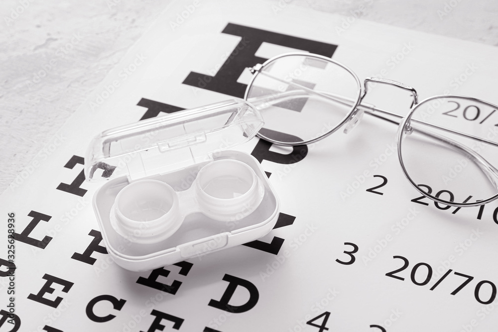 Container with contact lenses and glasses on eye test chart, closeup