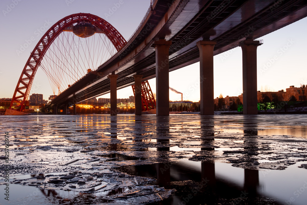 The photo shows picturesque bridge with big red arch over the river. This cable-stayed bridge stands