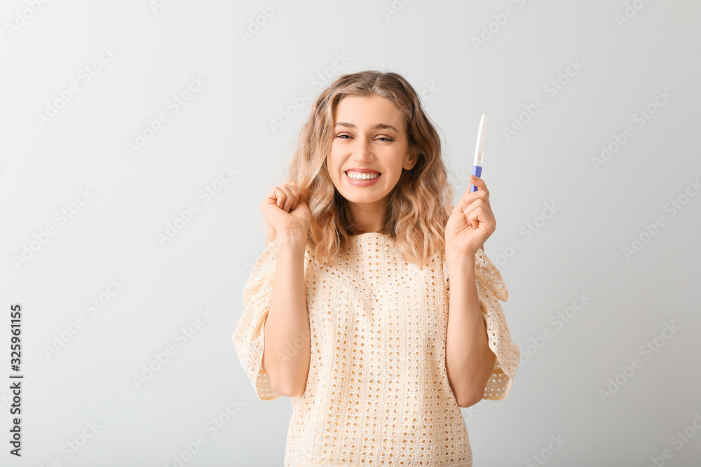 Happy young woman with pregnancy test on light background