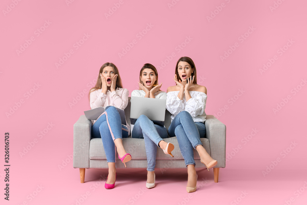 Surprised young women with laptop sitting on sofa against color background. Online shopping concept