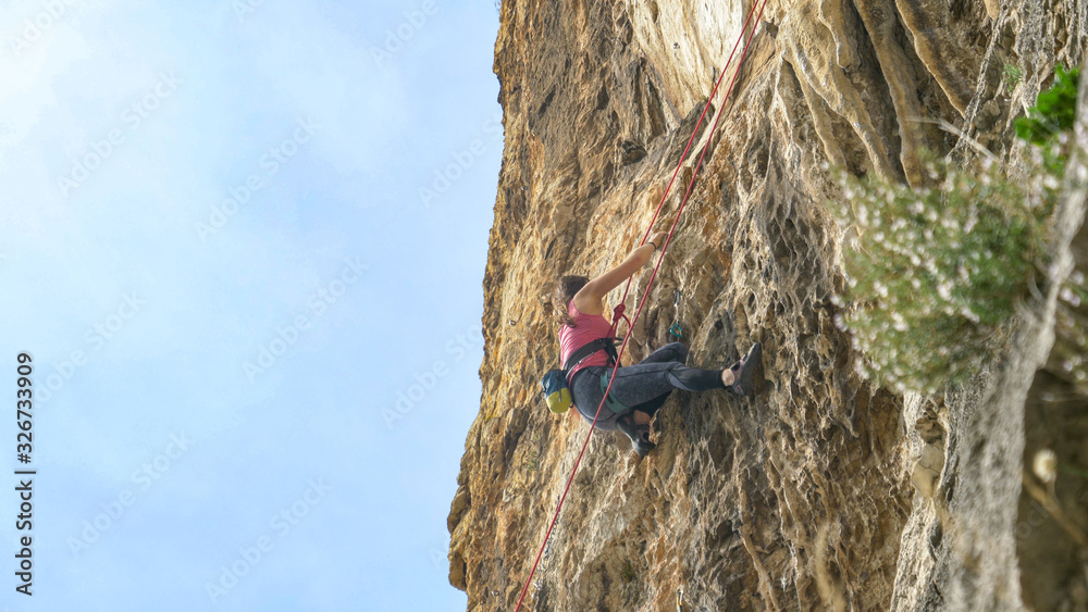 Young woman fearlessly climbs up a rocky cliff on perfect day for rock climbing