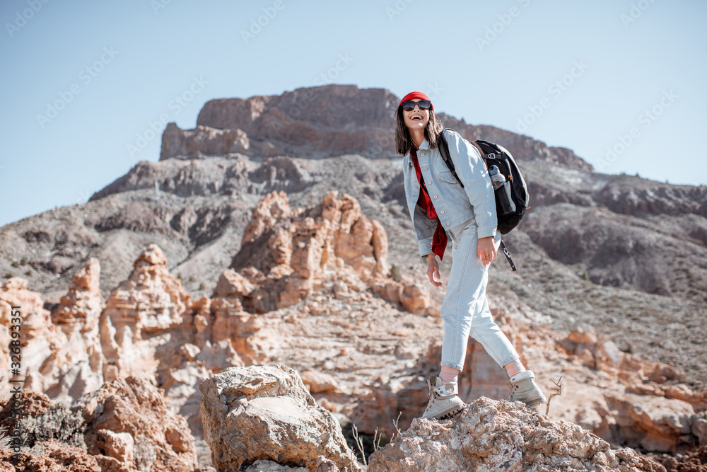 年轻女子穿着休闲牛仔裤，戴着红色帽子，背着背包，在阳光下在岩石地形上徒步旅行