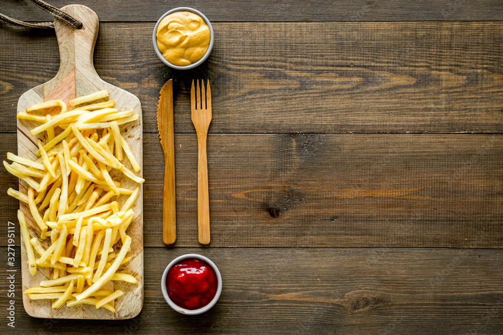 Fast food restaurant concept. French fries on cutting board near sauces on wooden table top-down cop