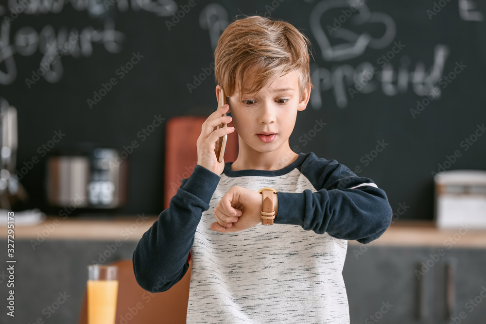 Surprised little boy looking at his watch while talking by phone in kitchen