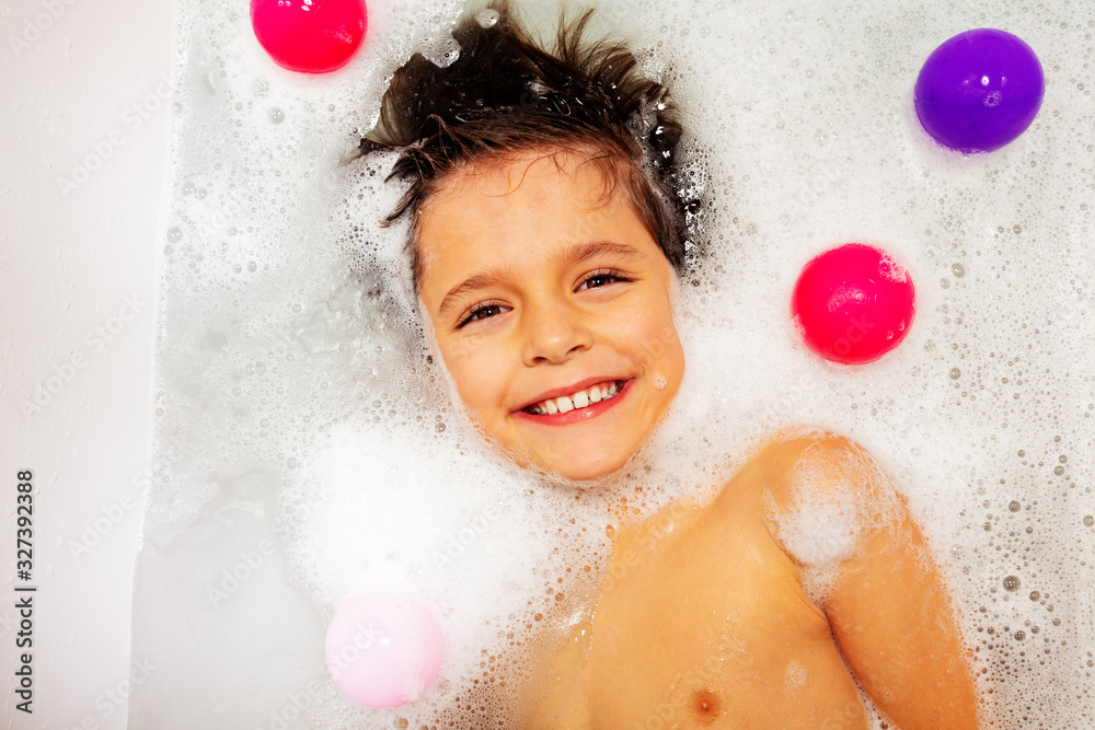 Portrait of cute boy with big smile swim on the back in soap at home bath submerged