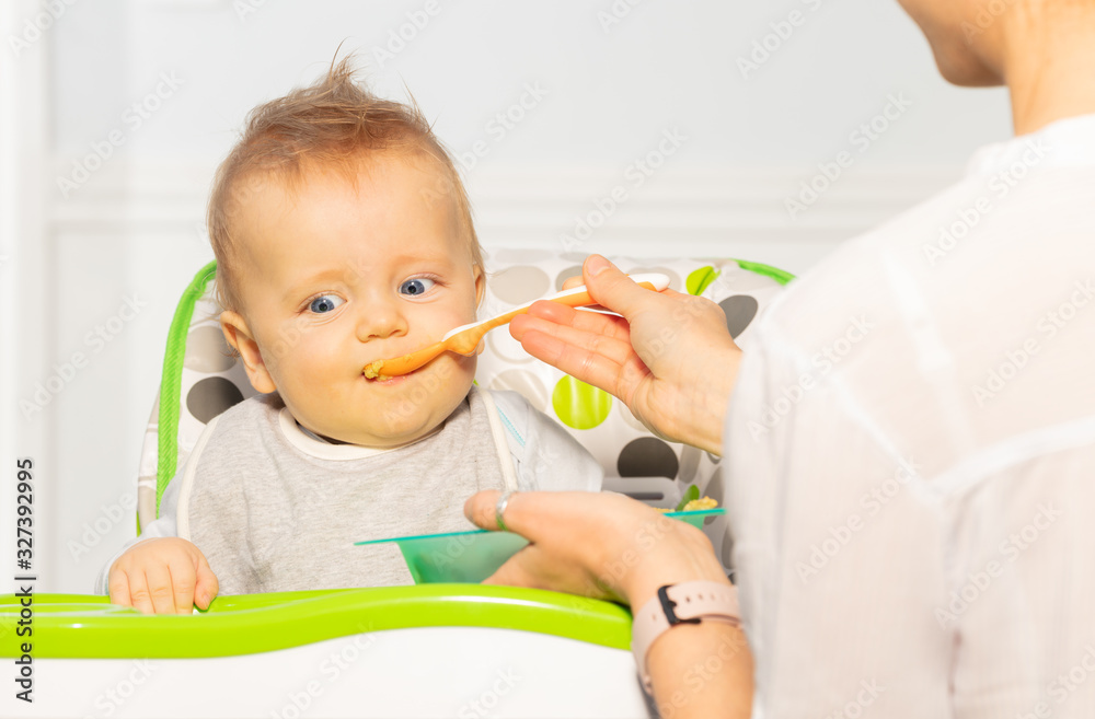 Mother feed little toddler baby boy with open mouth from plastic spoon in high chair view from back