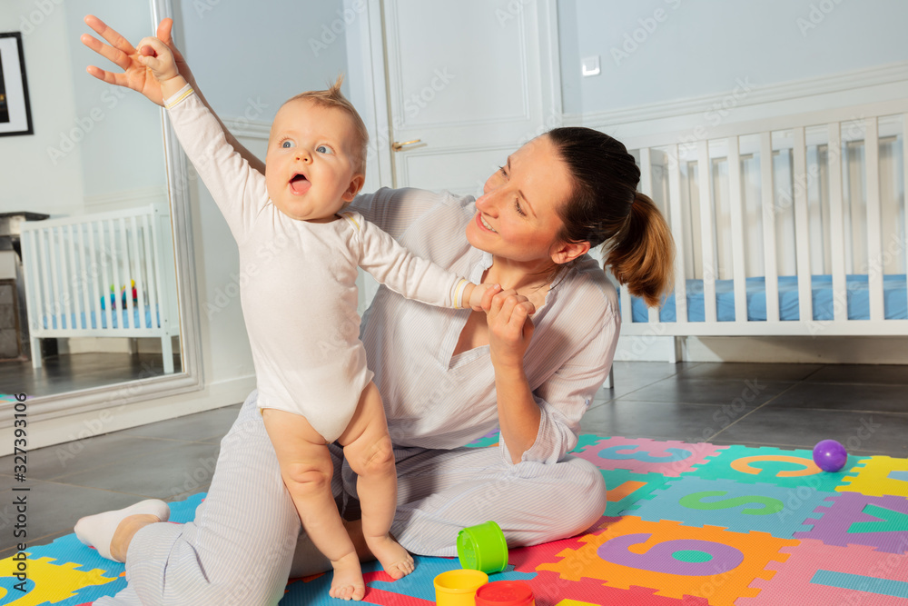 Happy mother play with toddler baby boy in the nursery helping him to stand and lift hands