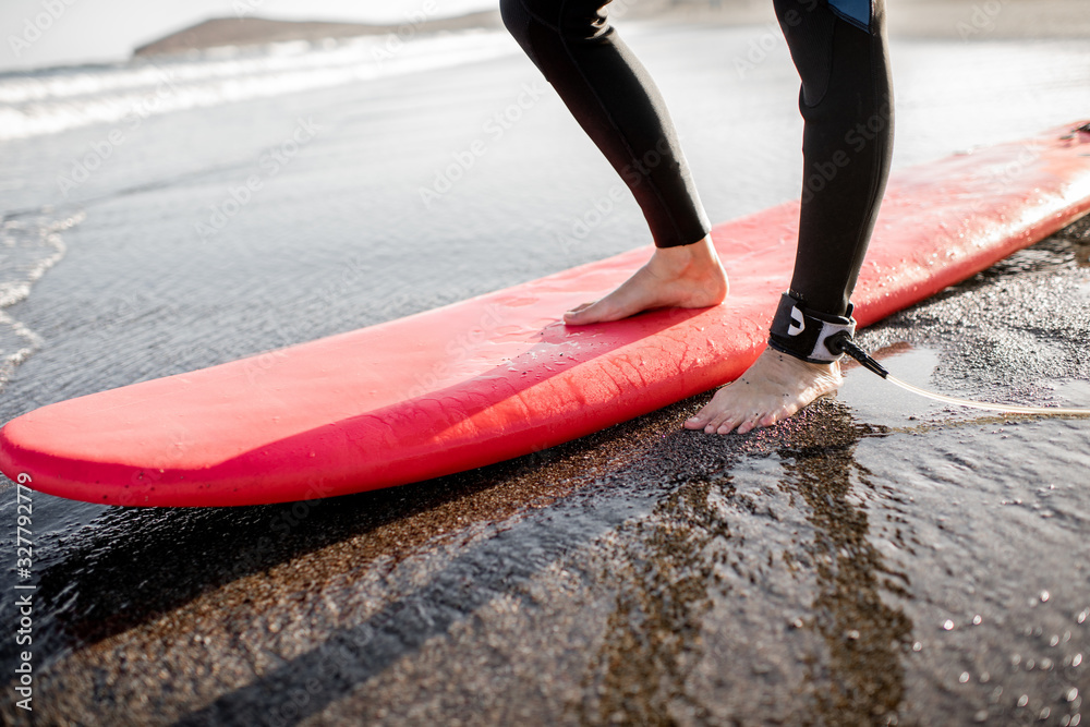 Surfer standing with surfboard on the sandy beach on a sunset, cropped view focused on womans legs