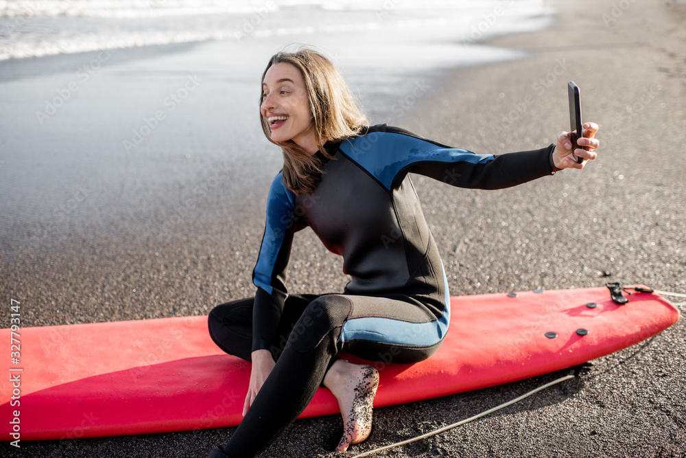 Young surfer in wetsuit making self portrait on phone while sitting with surfboard on the beach. Wat