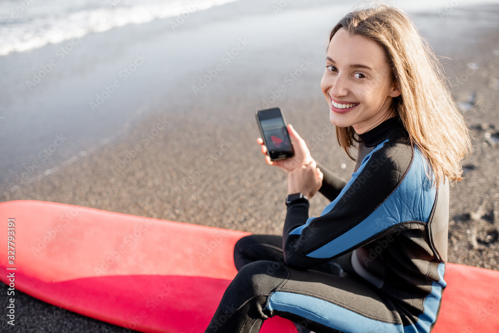 Portrait of a young relaxed surfer in wetsuit sitting with surfboard and smart phone on the beach. W