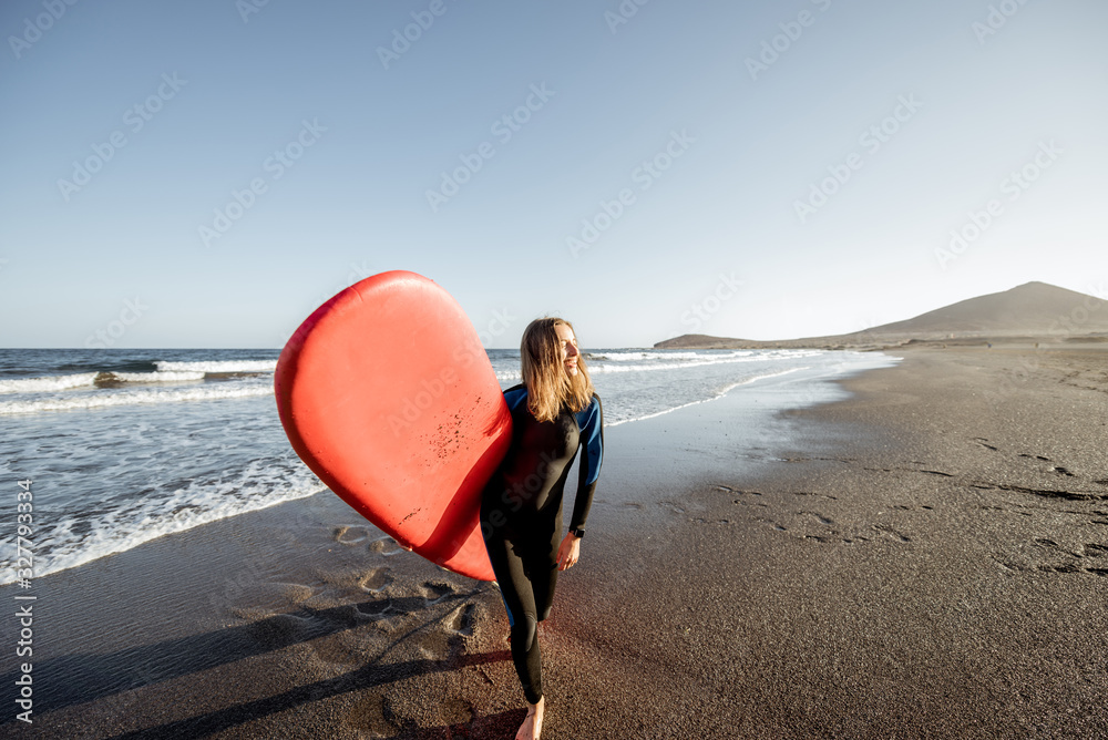 Young surfer in wetsuit walking with red surfboard on the beautiful ocean beach during a sunset, wid