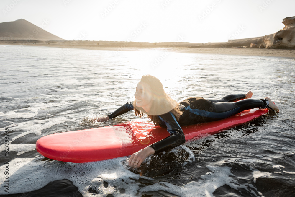 Young woman in wetsuit catching water flow on the surfboard, surfing on the wavy ocean during a suns