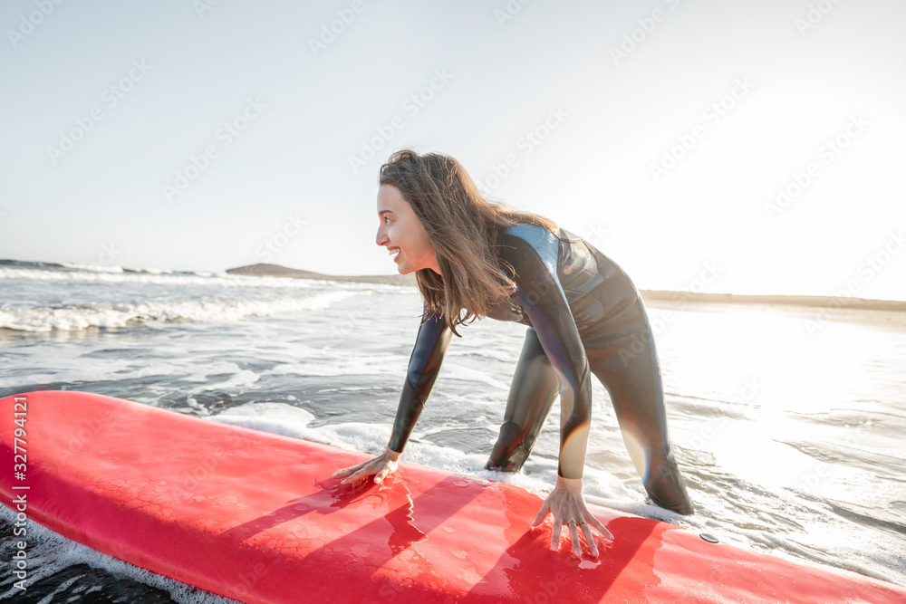 Young surfer in wetsuit getting on surfboard, catching water flow near the beach during a sunset. Wa