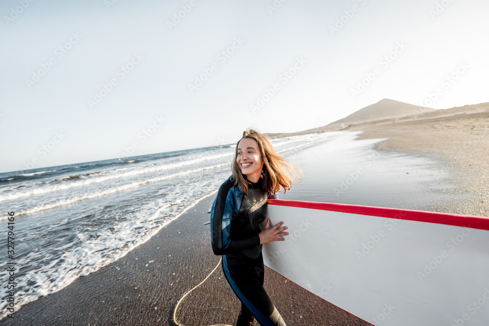 Portrait of a young joyful woman in wetsuit carrying surfboard on the ocean beach during a sunset. W