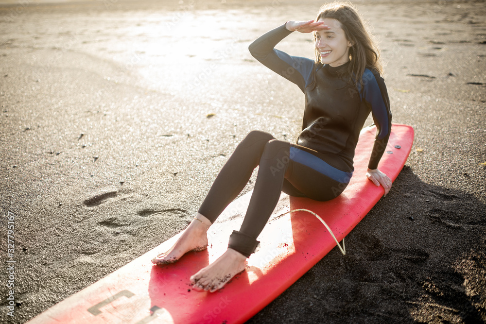 Portrait of a young relaxed surfer in wetsuit sitting with surfboard on the beach. Water sport and a