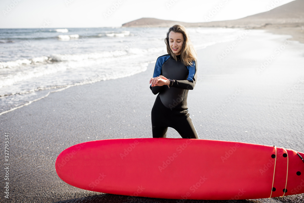 Young surfer in wetsuit standing with surf board on the wild beach on a sunset, looking on smart wat