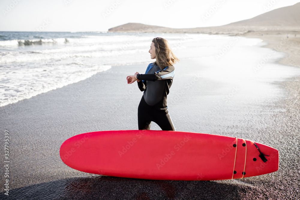 Young surfer in wetsuit standing with surf board on the wild beach on a sunset, looking on smart wat