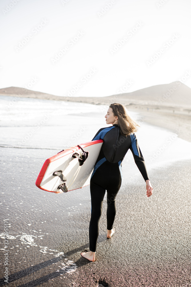 Young woman in wetsuit walking with surfboard, leaving footprints on the sand behind, view from the 