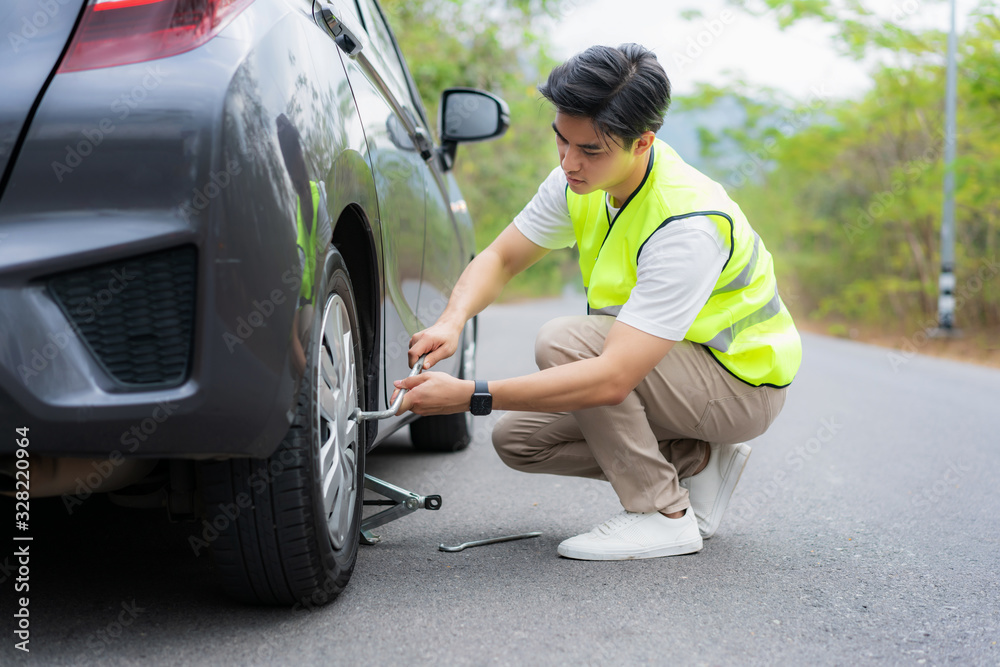 Young Asian man with green safety vest changing the punctured tyre on his car loosening the nuts wit