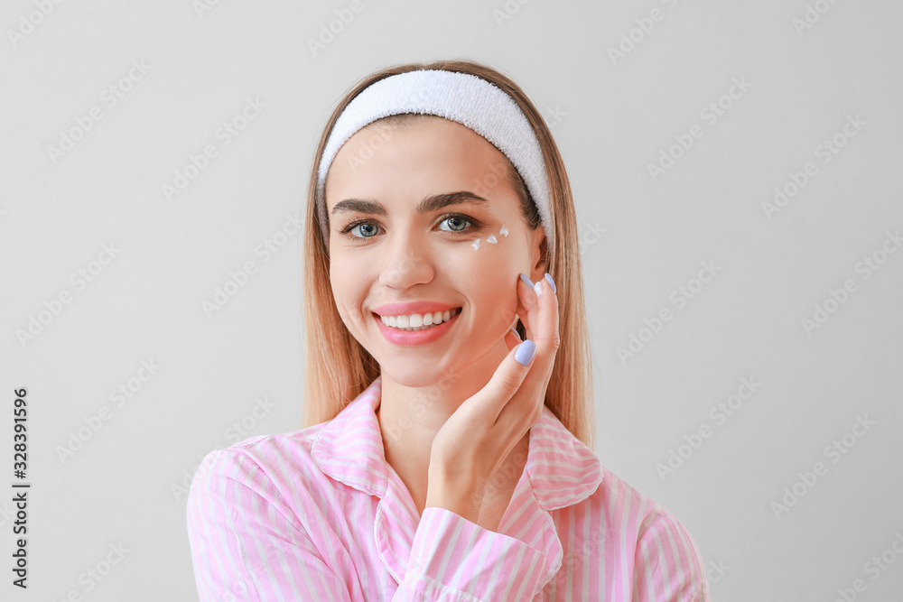 Beautiful young woman applying cream on her face against light background