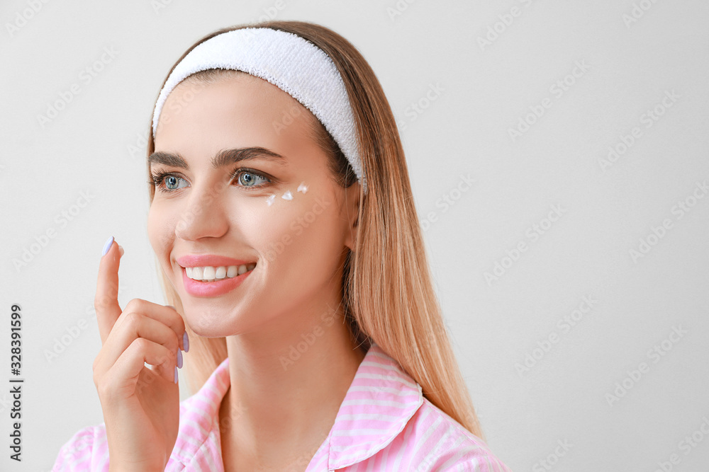 Beautiful young woman applying cream on her face against light background