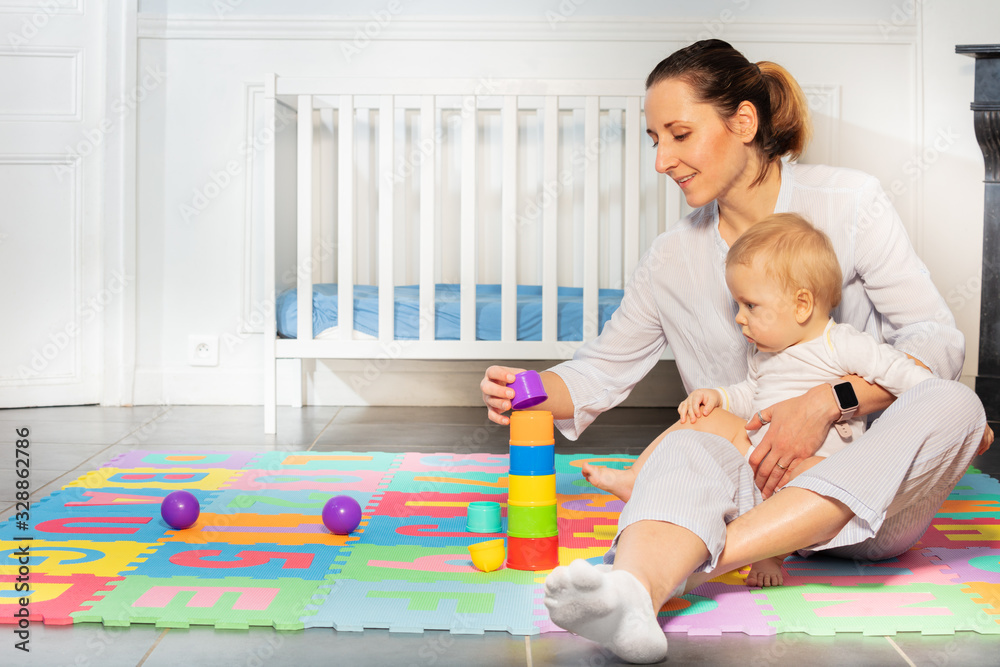 Mother play with little toddler baby boy sitting on children carpet near crib building pyramid of bl