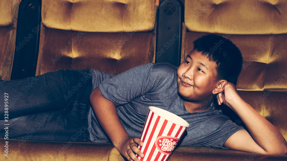 The boy is laying down watching movies and eating popcorn happily and enjoying in the cinema.