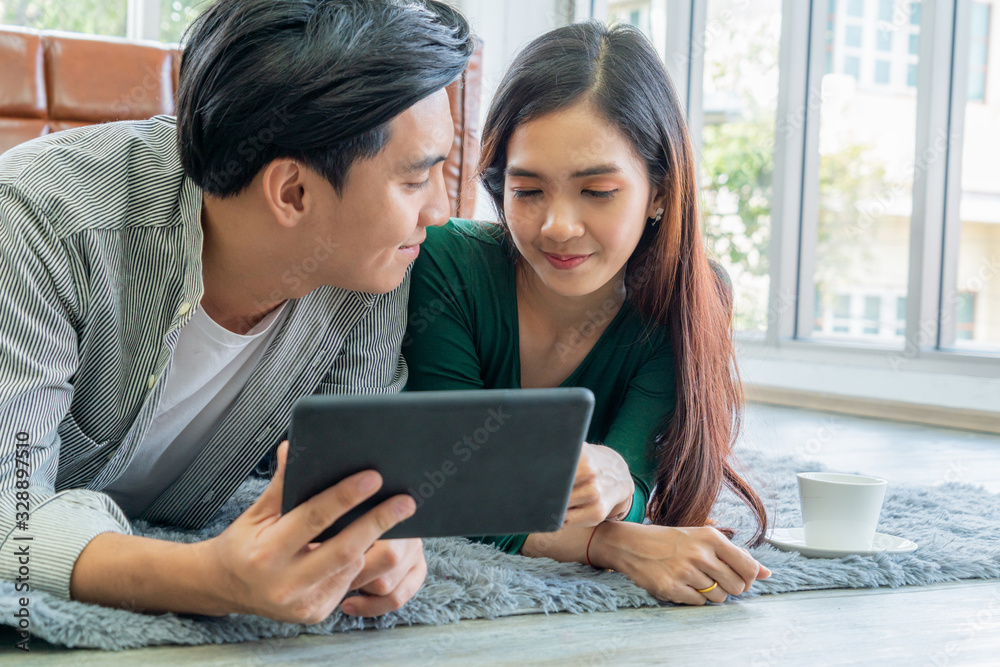 Happy Asian couple use tablet while lying down on carpet at living room floor. Love and relationship