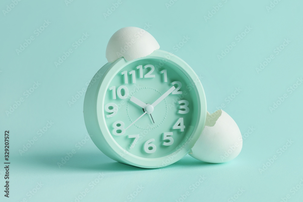 Alarm clock with broken egg shell on color background