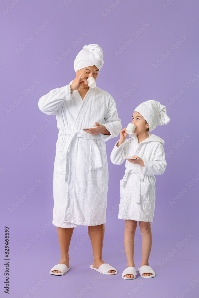 Asian man with his little daughter in bathrobes drinking tea on color background