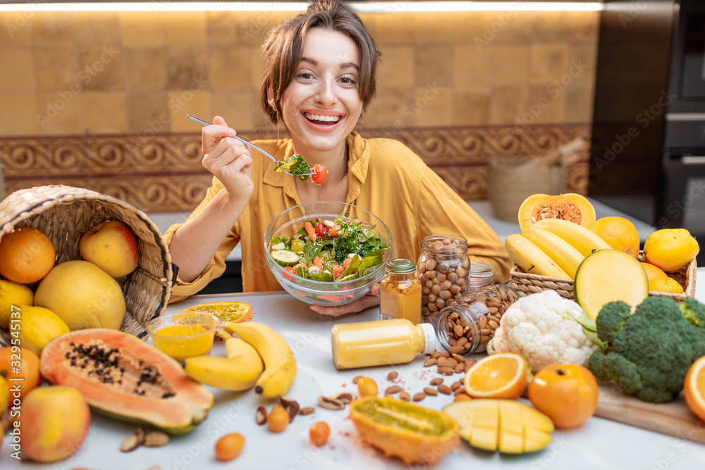 Portrait of a young cheerful woman eating salad at the table full of healthy raw vegetables and frui