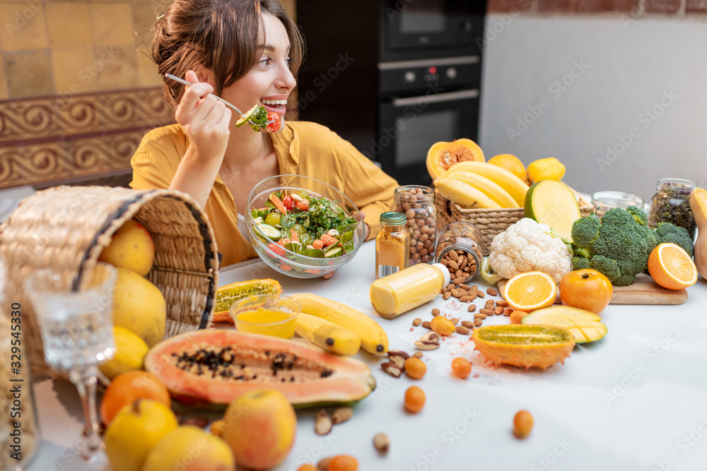 Portrait of a young cheerful woman eating salad at the table full of healthy raw vegetables and frui