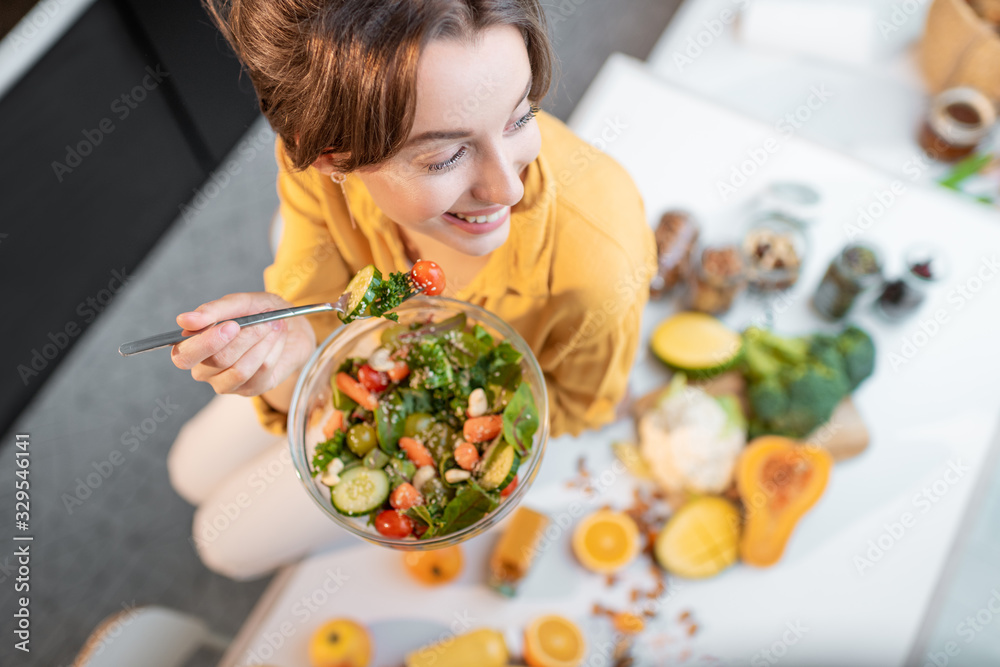 Portrait of a young cheerful woman eating salad at the table full of healthy raw vegetables and frui